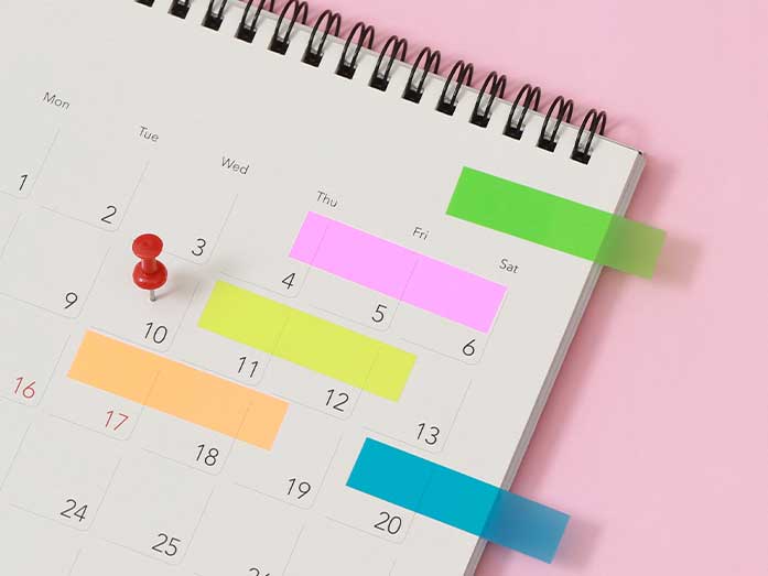 Open calendar with coloured sticky notes marking different dates and a red push pin on one day, set on a pink background.
